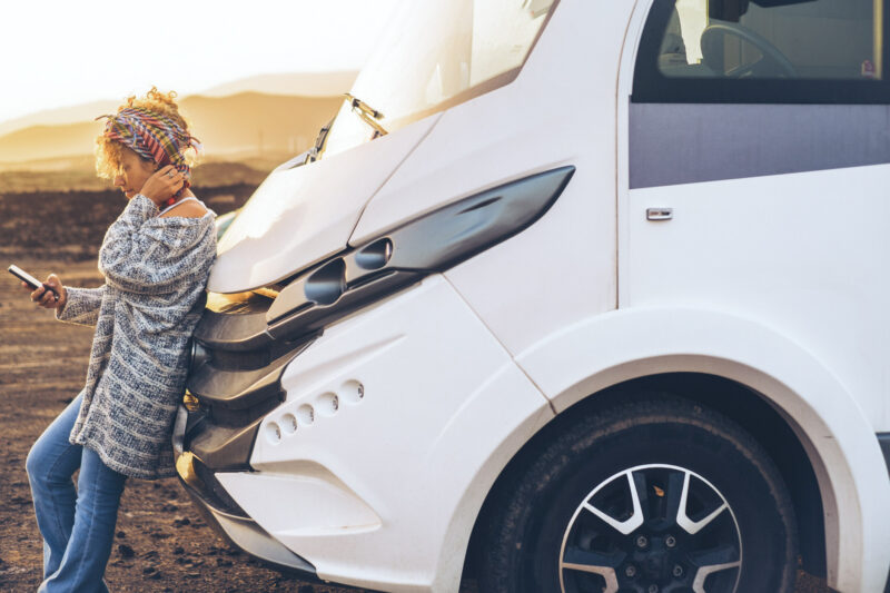 Travel people lifestyle van life with modern camper van. Adult young woman enjoy freedom against a motor home vehicle using mobile phone to plan next destination road trip. Female independent